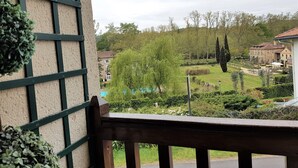 A view from a balcony with a railing, overlooking a well-maintained garden with various plants and a distant building with a tiled roof.