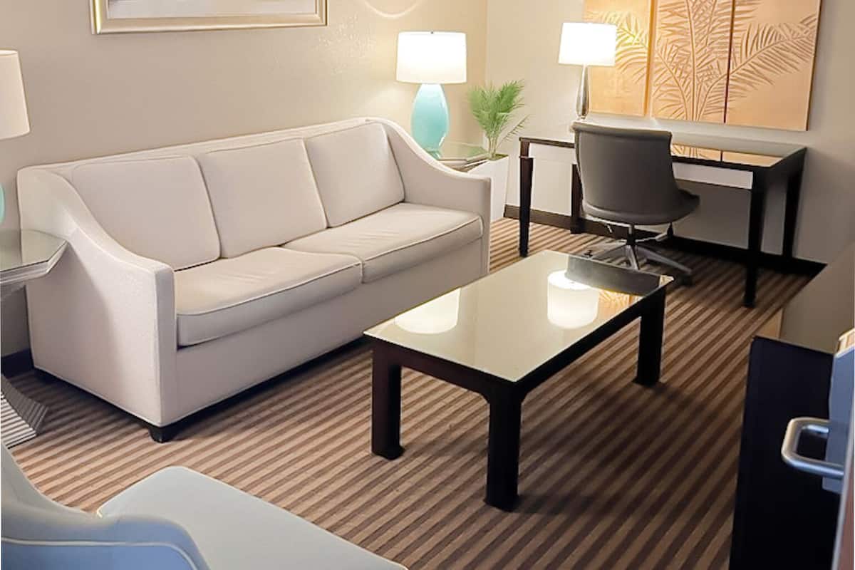 Habitación de hotel con un sofá blanco, una mesa de centro de vidrio, un sillón azul, un escritorio con silla, una lámpara y una pared con diseño de hojas.