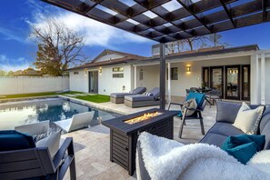 Een modern terras buiten met een zwembad, lounge stoelen en een vuurkuil.
