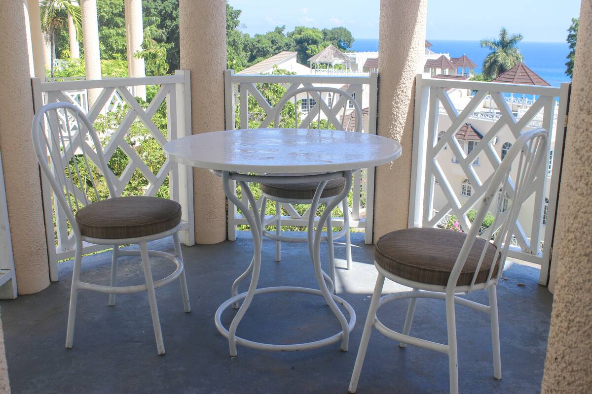 A balcony with two white chairs and a small table, overlooking a scenic view of the ocean and buildings.