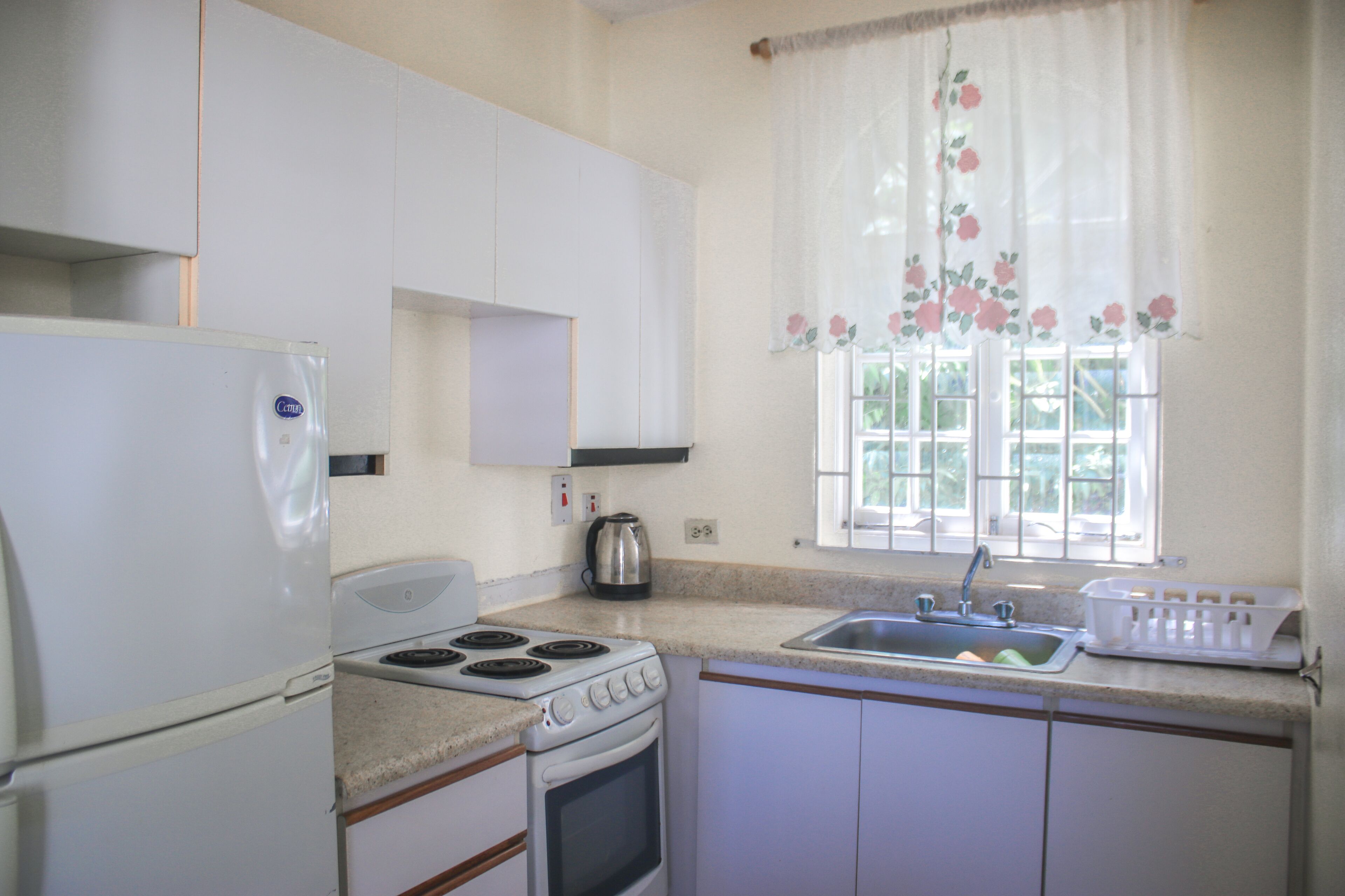 A kitchen with a refrigerator, stove, oven, sink, and window with floral curtains.
