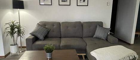 Een woonkamer met een grijze hoekbank, een houten salontafel, een kleine potplant en een vloerlamp. Er hangen drie ingelijste foto's aan de muur.