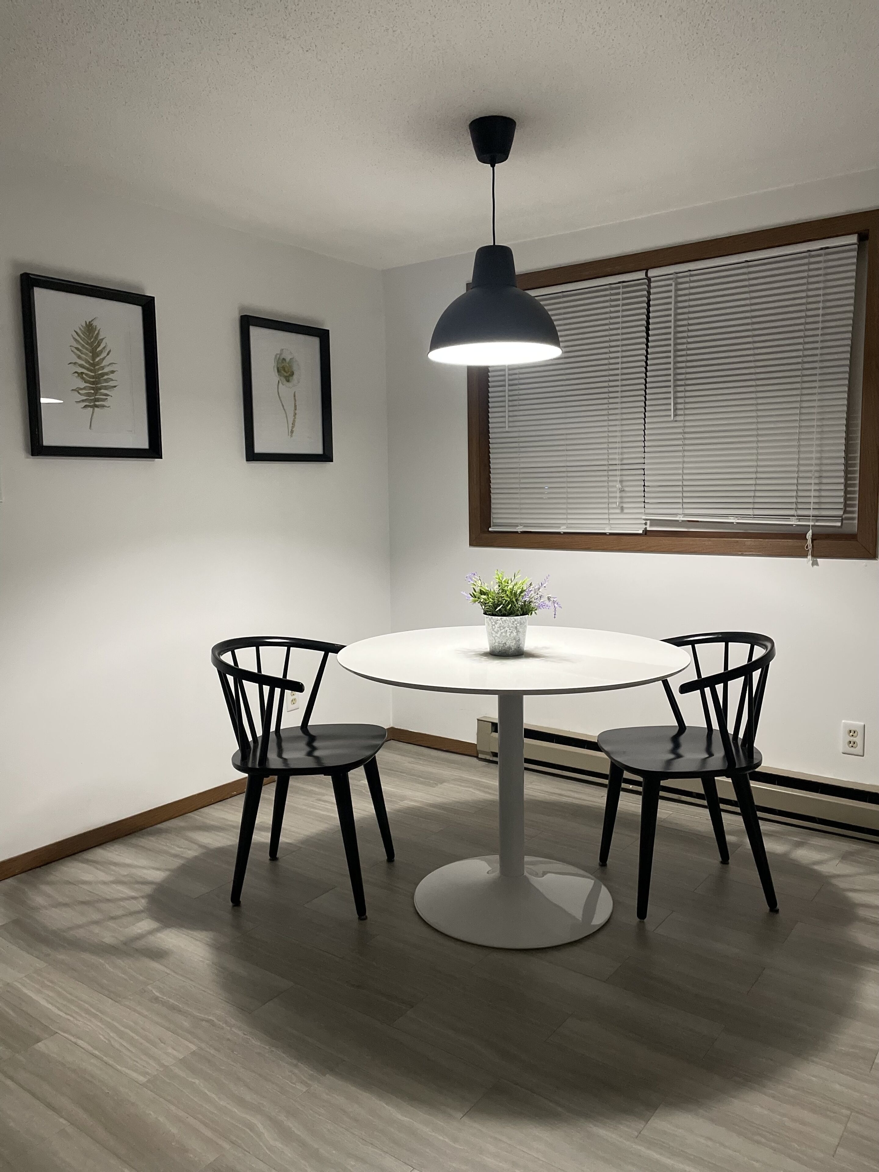 Un coin repas avec une table ronde, deux chaises, une suspension et des reproductions encadrées de motifs botaniques accrochées au mur.