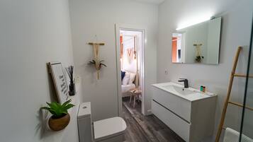 Un baño moderno con inodoro, lavabo y espejo. Hay una mesita de noche con una planta y una cita enmarcada en la pared.