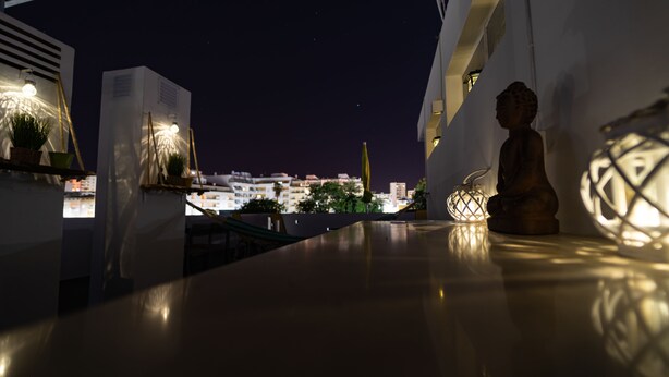 En tagterrasse med udsigt over bybilledet om natten, der inkluderer en statue, et bord og en hængende lampe.