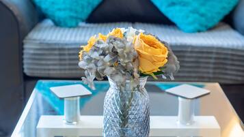 Una sala de estar moderna con una mesa de centro de vidrio, un florero con flores y un sofá gris con cojines color turquesa.