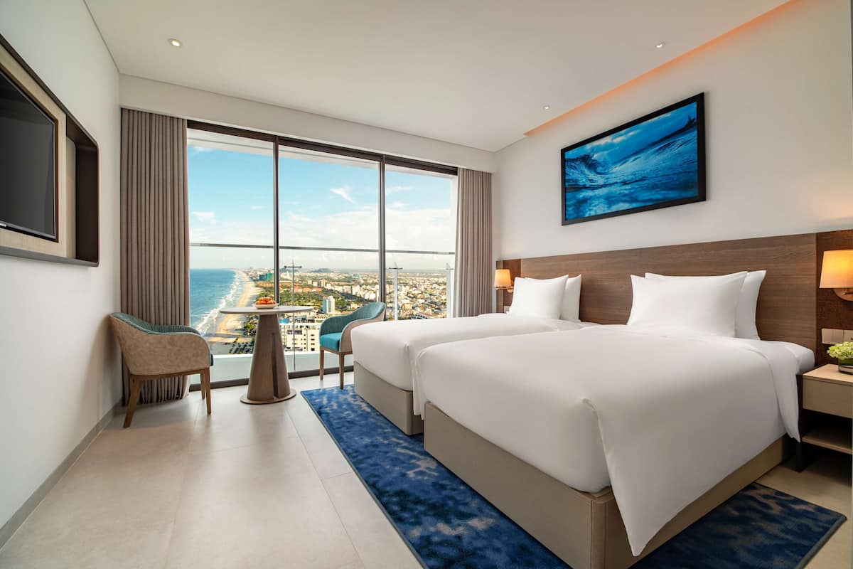 A hotel room with two beds, a TV, a large window with a city view, and a painting on the wall.