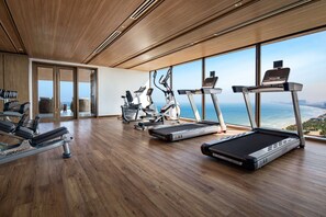 A modern gym with various cardio machines, large windows offering ocean views, and wooden flooring.