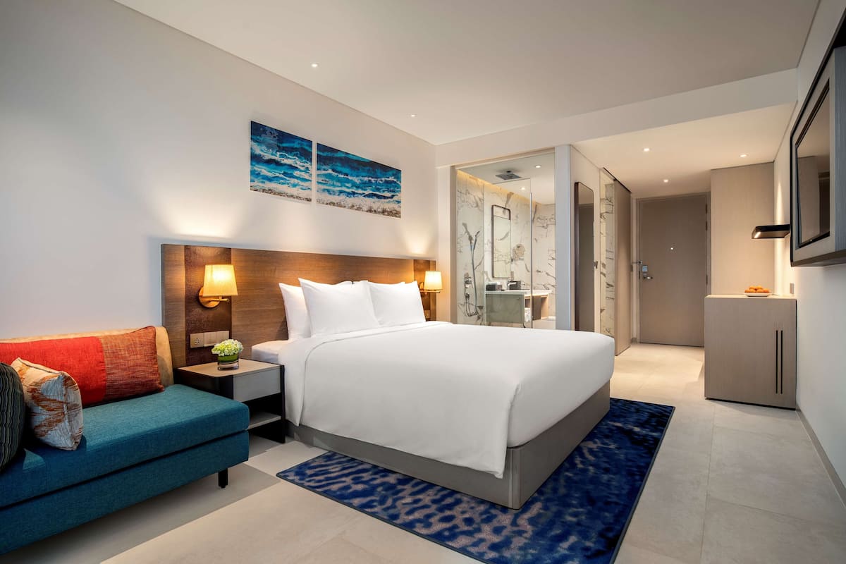 A modern hotel room with a large bed, a sofa, a desk, and a bathroom visible through an open door.