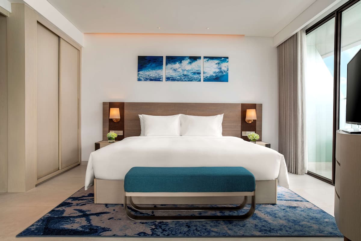 A modern hotel room with a large bed, a bench, a TV, and a view of the city.