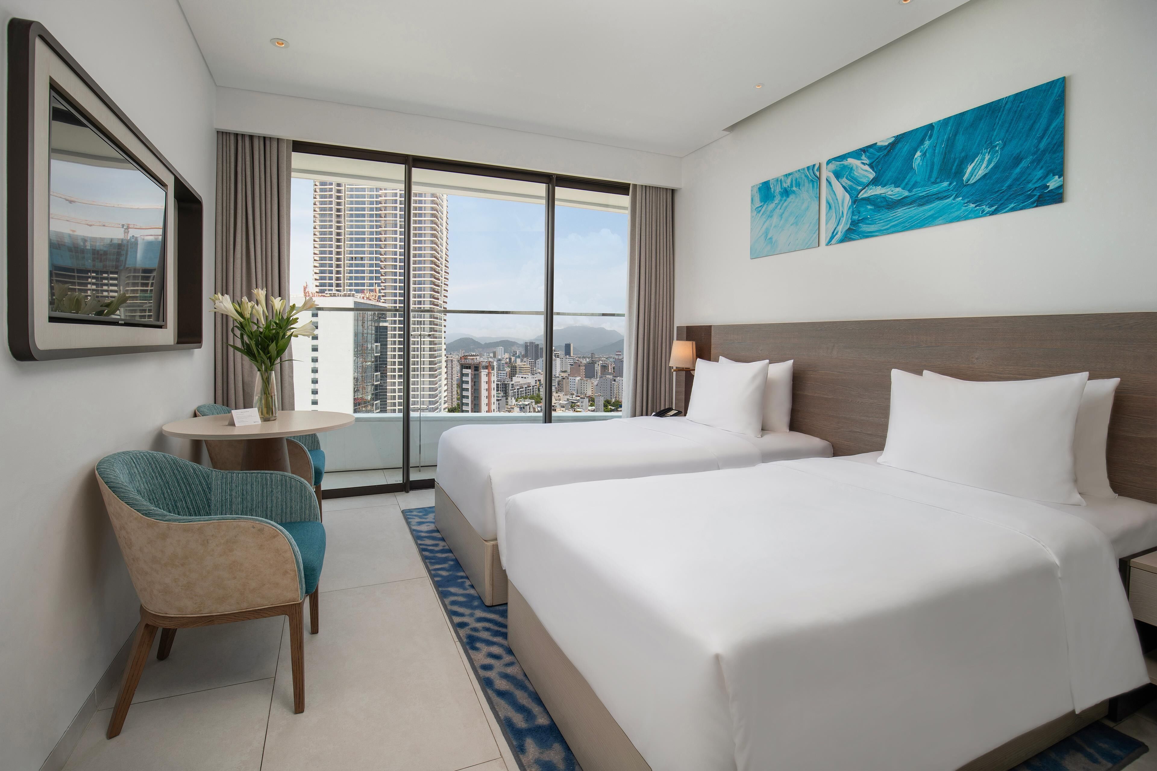 A hotel room with two beds, a view of the cityscape, a framed picture on the wall, and a small table with a vase of flowers.