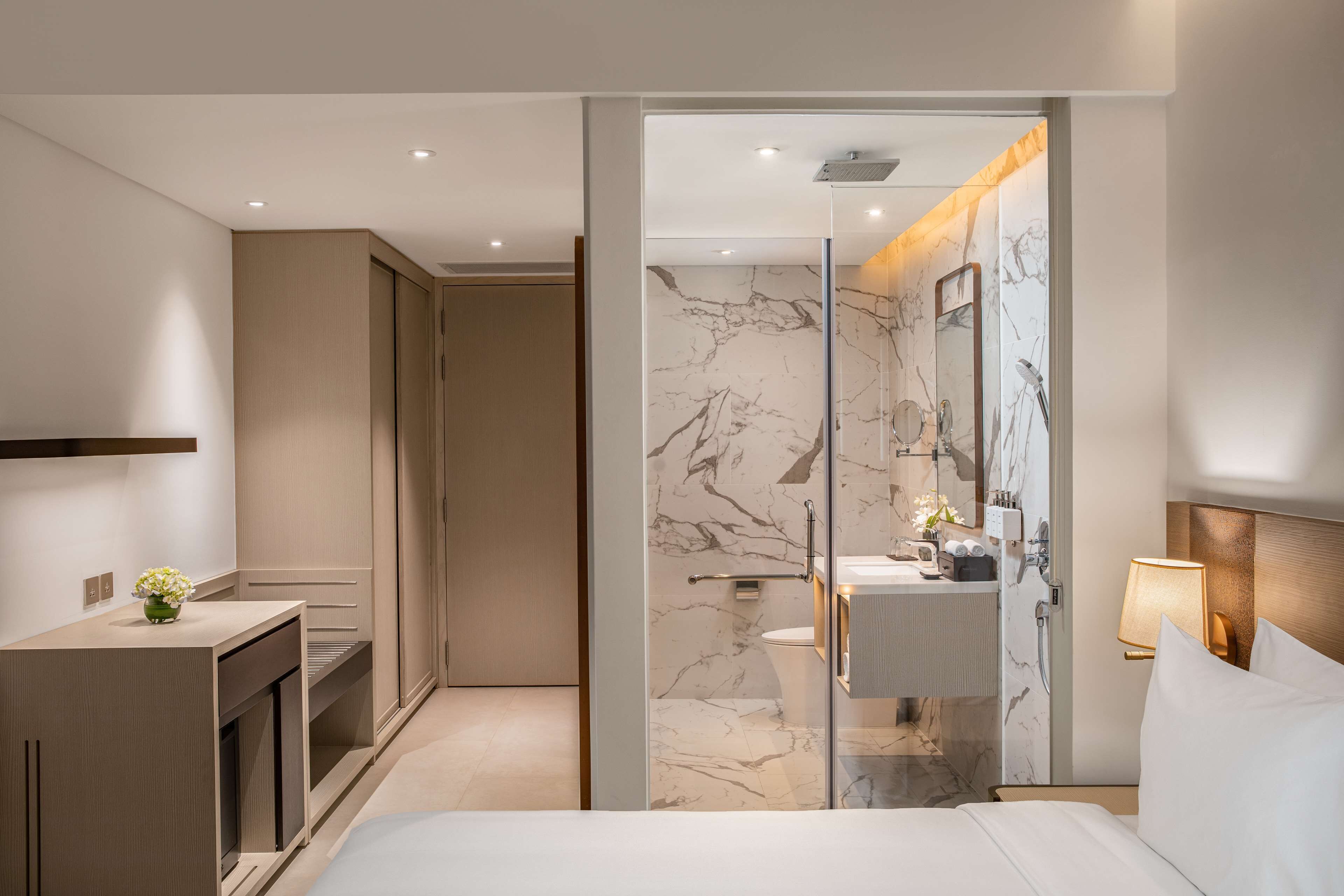 Una habitación de hotel moderna con baño de vidrio, una cama, una mesita de noche y otra mesita auxiliar.