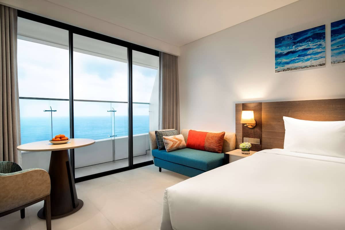 A hotel room with a bed, a sofa, a table with a bowl of fruit, a nightstand with a lamp, and a large window with a view of the sea.