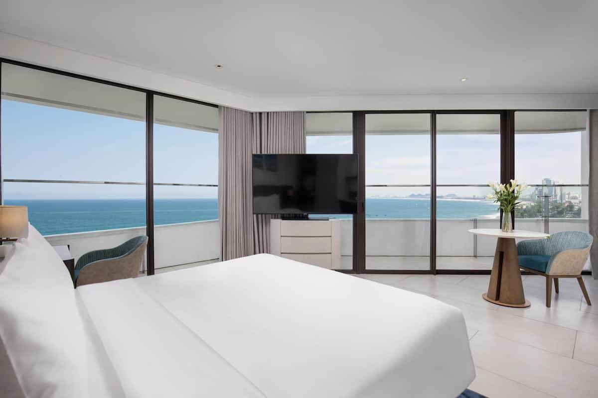 A modern hotel room with a large bed, a TV, a small table, and a chair. The room has large windows offering a view of the ocean.