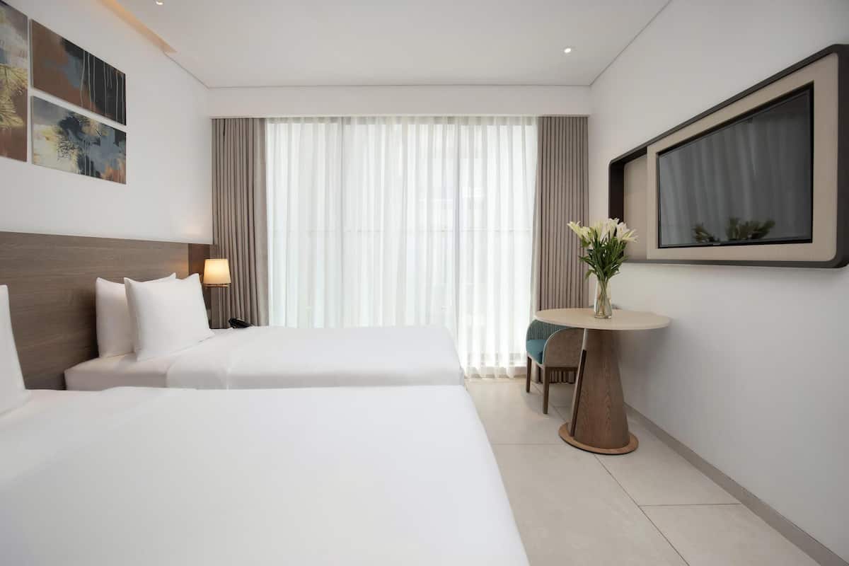A hotel room with two beds, a round table with a vase of flowers, and a large window with curtains.