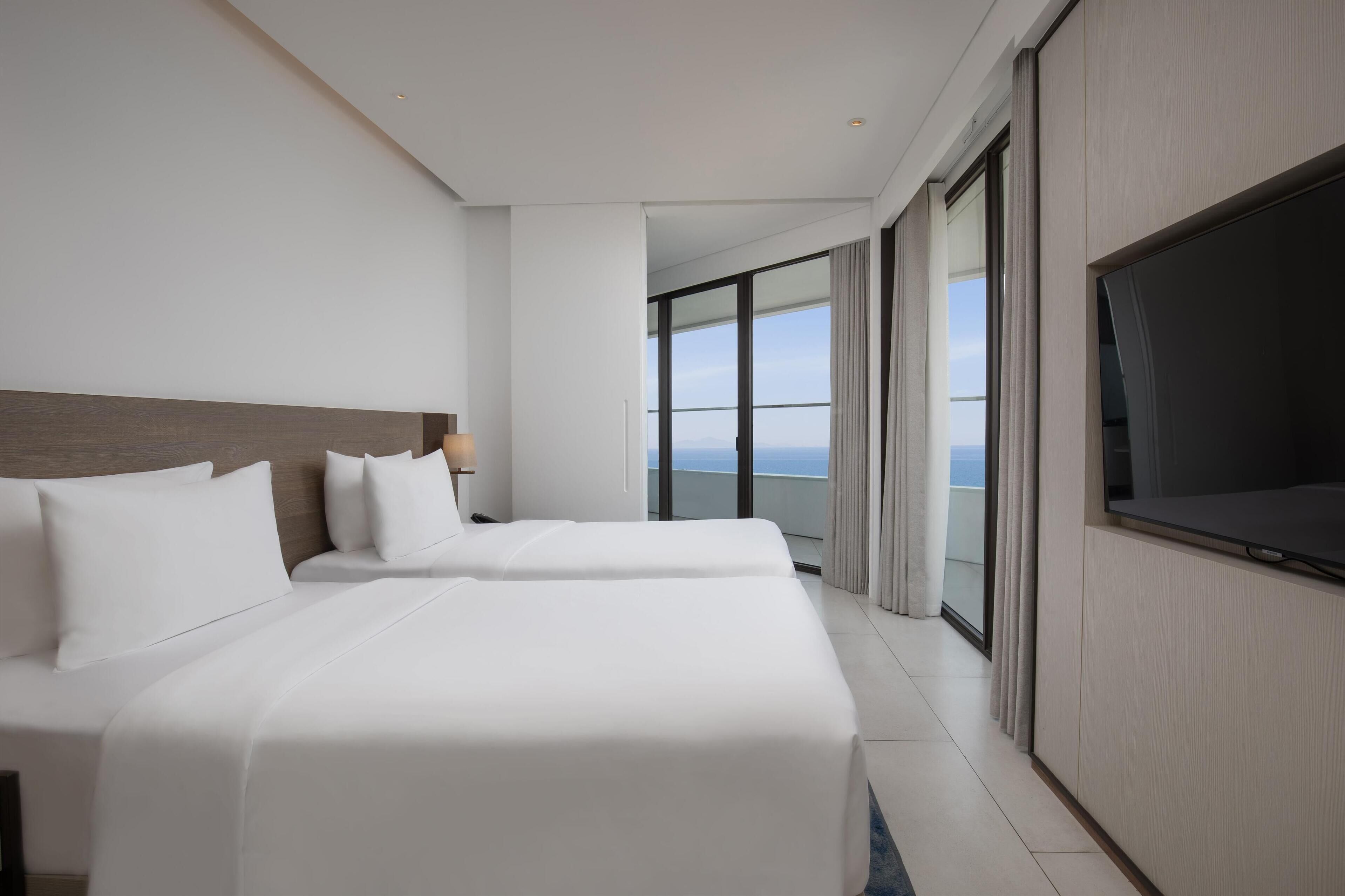 A hotel room with two beds, a large TV, and a balcony with a view of the sea.