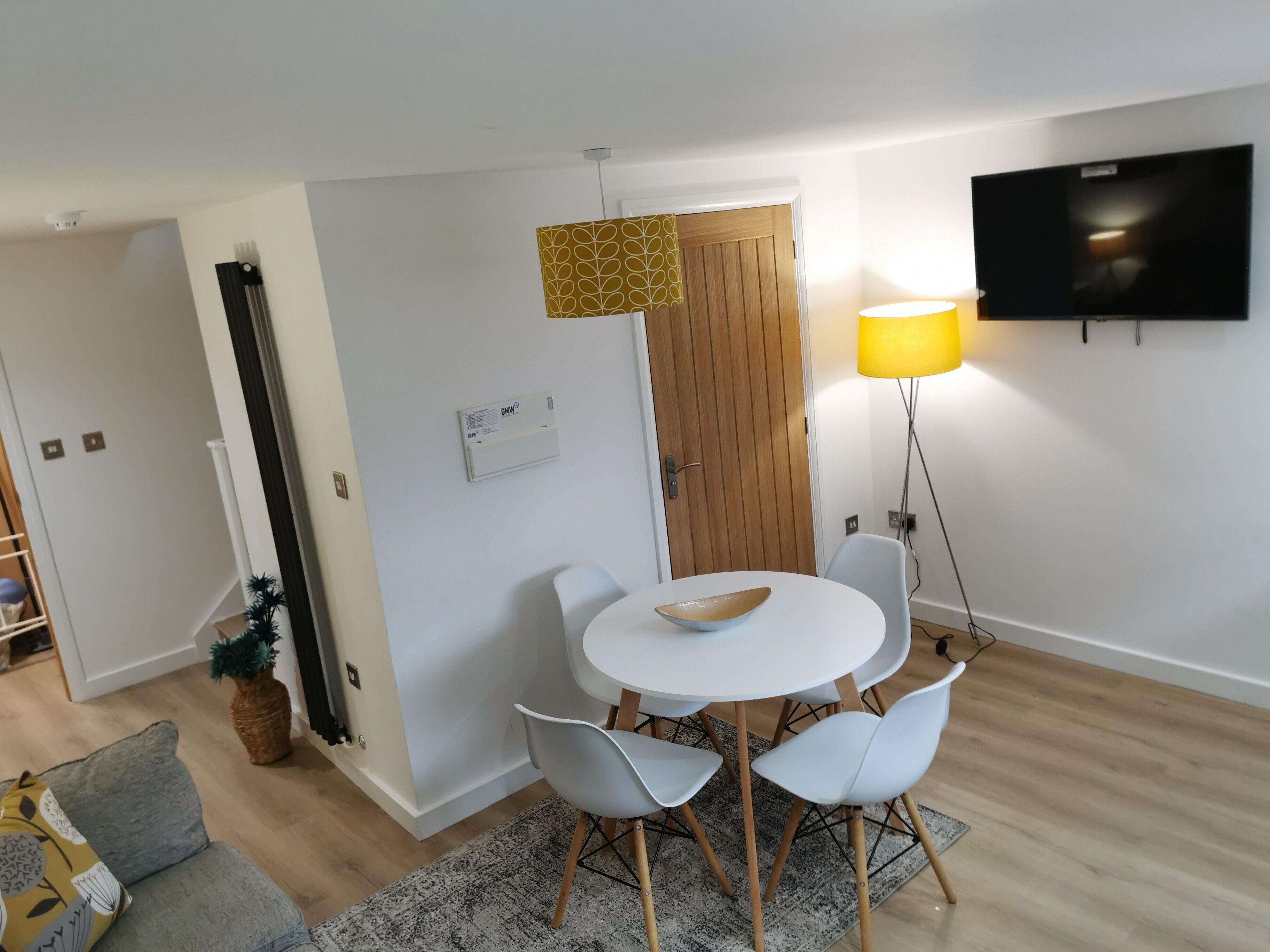 A modern living room with a round white table, four white chairs, a wooden door, a mounted TV, and a floor lamp.