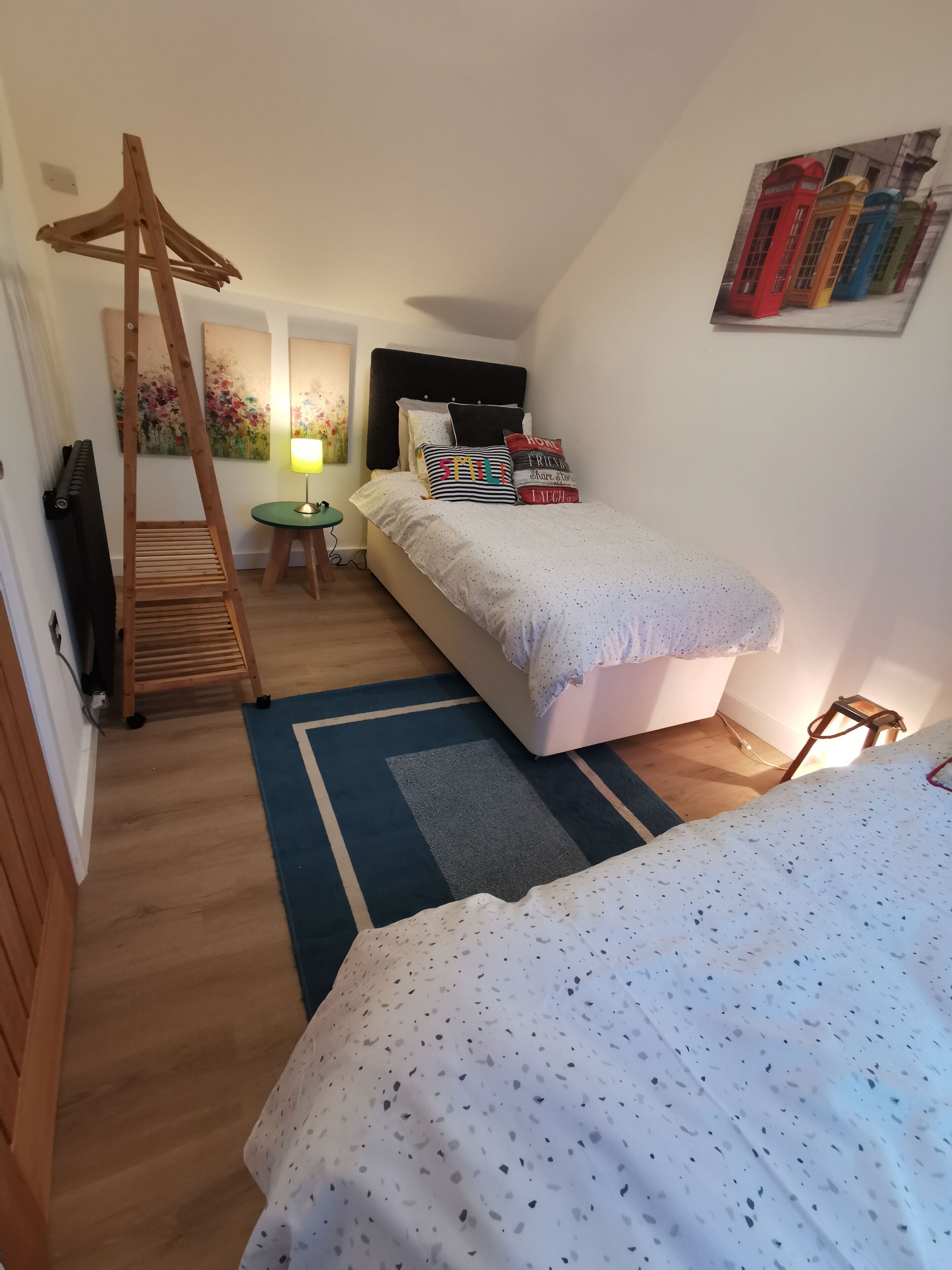 A bedroom with a bed, a wooden ladder, a small table with a lamp, and a painting of red telephone booths.