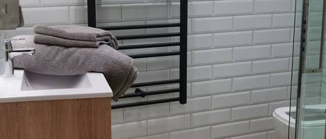 Un baño moderno con pared de azulejos blancos, un toallero negro, un tocador de madera y una cabina de ducha de vidrio.