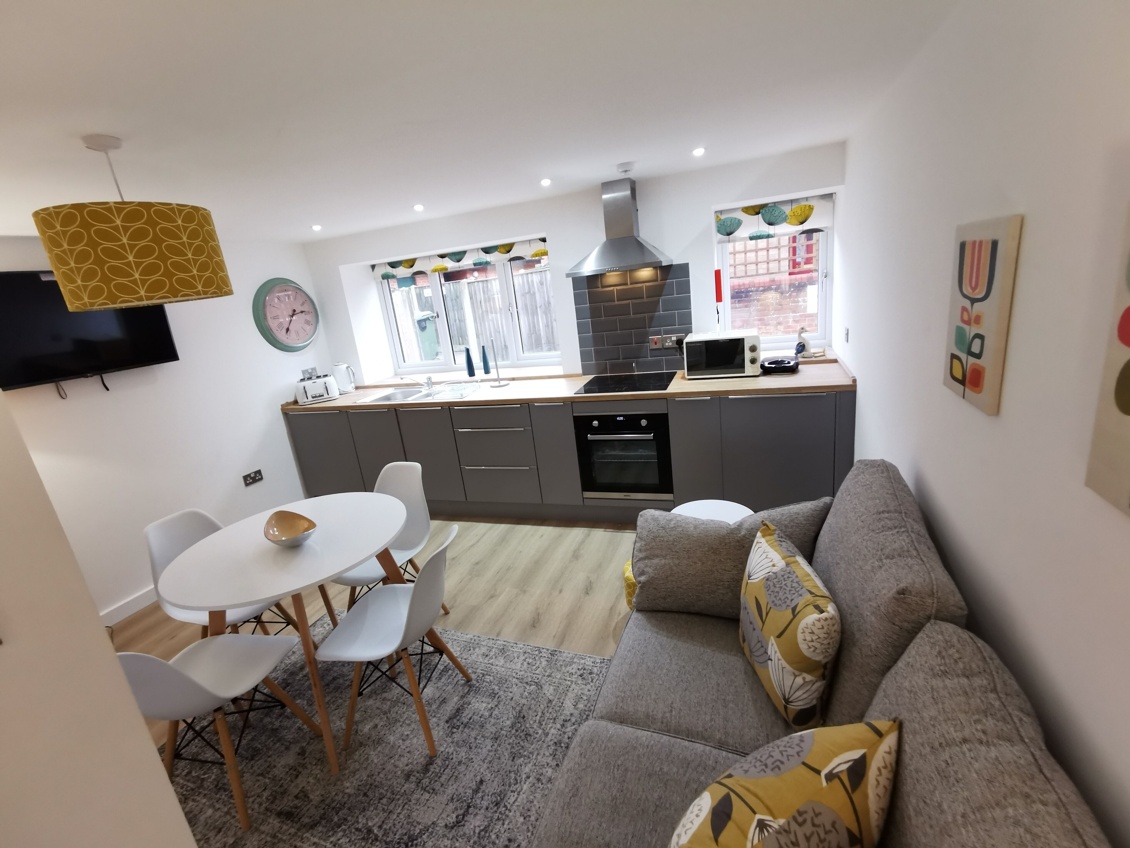 A modern kitchen with a built-in oven, a microwave, and a large window. There is a dining area with a white table and chairs, and a grey sofa with patterned cushions.