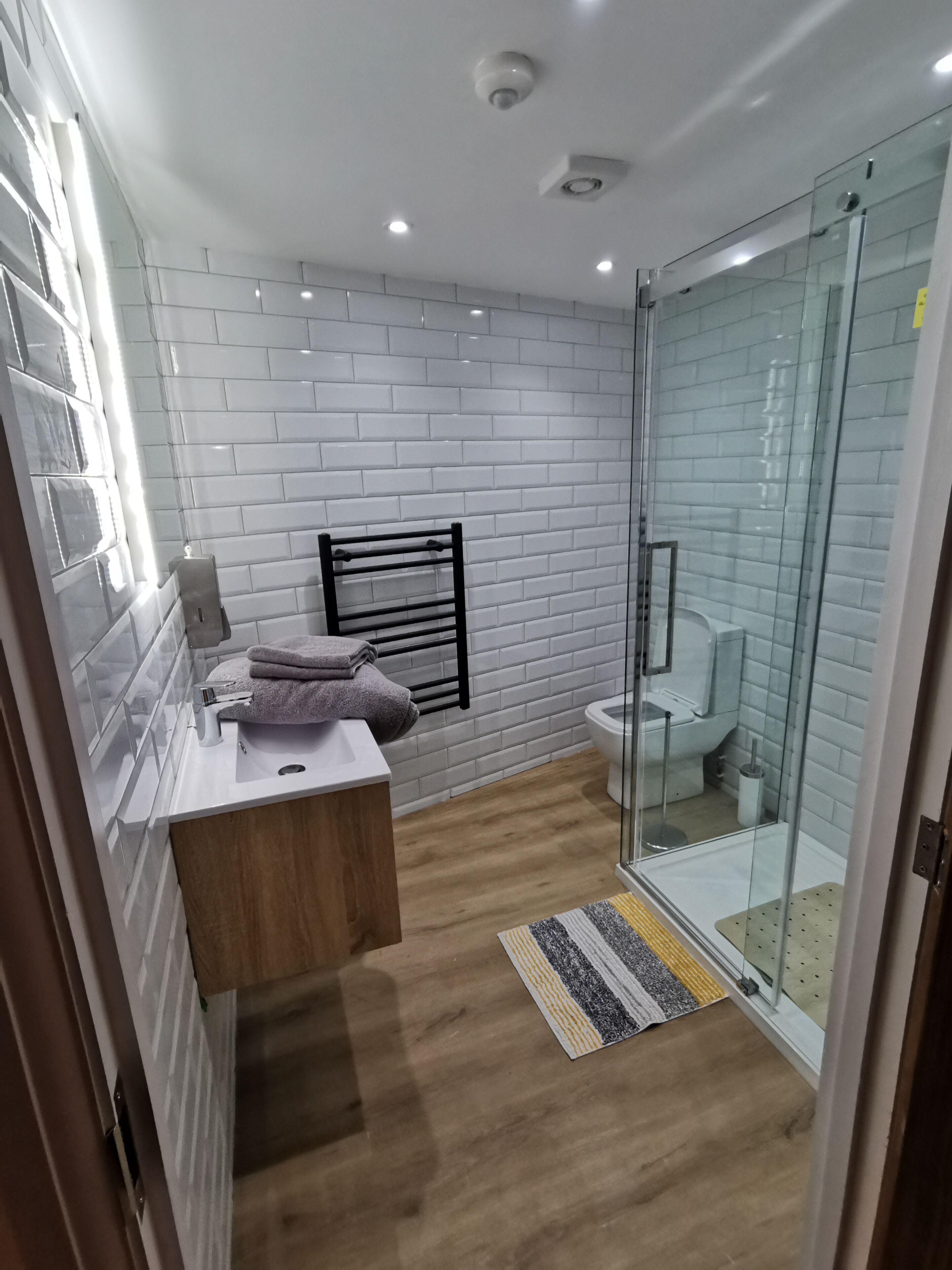 A modern bathroom with white tiled walls, a wooden vanity, a glass shower enclosure, and a toilet.