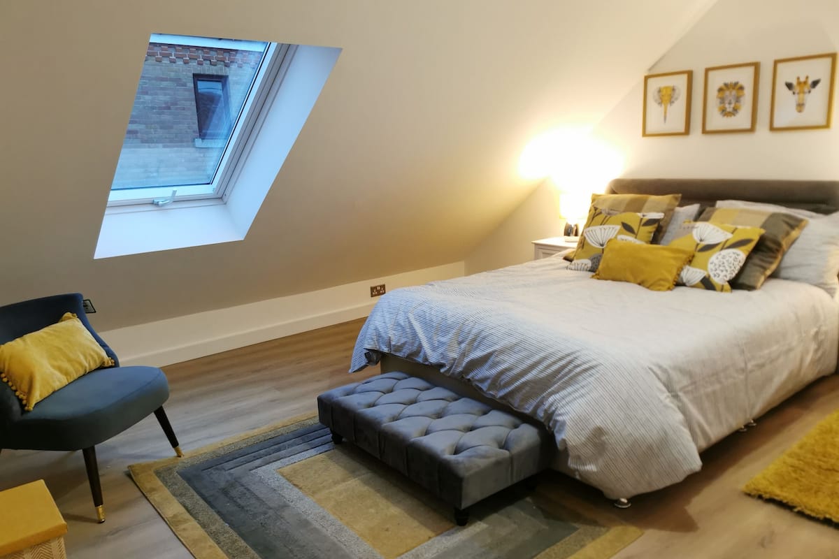 A bedroom with a loft-style ceiling, a bed with multiple pillows, a chair, a small table, and a window with a view of the outside.