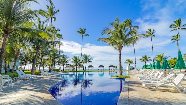 A pool area with lounge chairs and umbrellas, surrounded by palm trees and a clear sky. - Ancorar mar-piscina em Porto de Galinhas (Ipojuca)