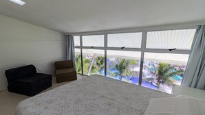 A bedroom with a large bed, a dark armchair, a brown sofa, and a view of palm trees through large windows. - Ancorar mar-piscina em Porto de Galinhas (Ipojuca)