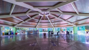 A spacious indoor area with a high ceiling featuring wooden beams and a central light fixture. There are several tables and chairs arranged for dining or socializing. - Ancorar mar-piscina em Porto de Galinhas (Ipojuca)