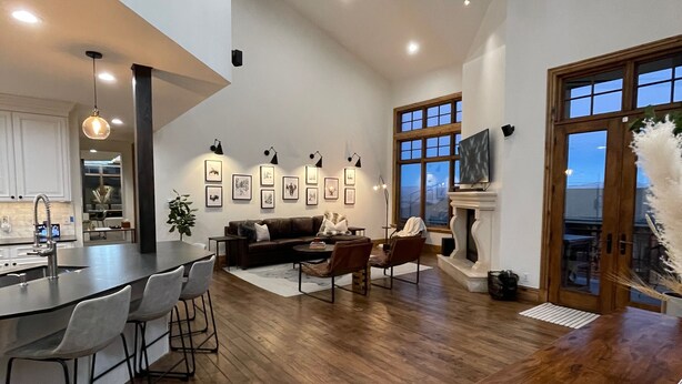 Een ruime woonkamer met houten vloer, een eethoek met barstoelen, een open haard en een groot raam.