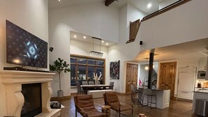 Een ruime woonkamer met een open haard, een grote flatscreen-tv, leren banken en een eethoek, met een keuken op de achtergrond.