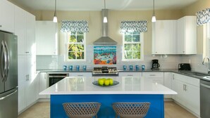Een moderne keuken met witte kasten, een centraal kookeiland met barkrukken, een roestvrijstalen koelkast en een kleurrijke spatwand.