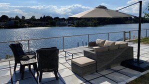 A terrace with wicker furniture, a table, and a view of a lake with houses in the background.