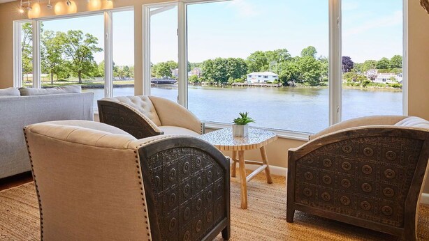 A room with a large window overlooking a lake, a sofa, two armchairs, and a small table with a plant.