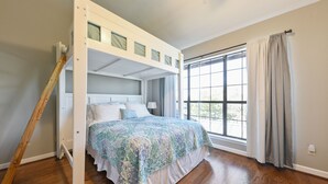 A bedroom with a bunk bed, wooden flooring, a large window with curtains, and a view of the outdoors. - Comal River Getaway at the Froh Haus C206 (New Braunfels)