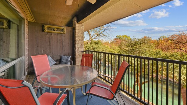 A covered outdoor patio with a round table and chairs, a ceiling fan, and a view of trees and a river. - Comal River Getaway at the Froh Haus C206 (New Braunfels)