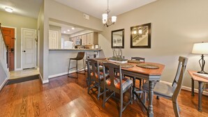 A dining area with a wooden table, chairs, and a chandelier. There is a kitchen in the background with cabinets and a microwave. - Comal River Getaway at the Froh Haus C206 (New Braunfels)