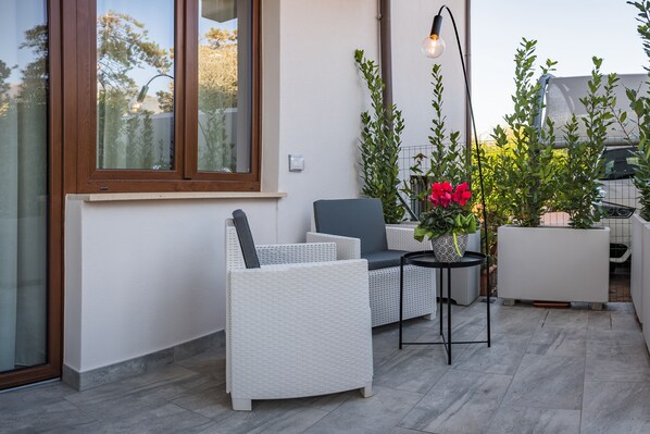 A patio with wicker furniture, a small table with a potted plant, and a window with a view of trees. - Beautiful Assisi Suite (Assisi)