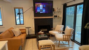 A living room with a stone fireplace, a flat-screen TV mounted above it, a sofa, two armchairs, and a coffee table. - Andora Villa #123 Newly Remodeled 2 BR / 2Bath, Centrally Located, Close to Town Center (Ketchum)