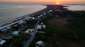An aerial view of a coastal area with a road running parallel to the beach, houses along the road, and a sunset over the ocean.