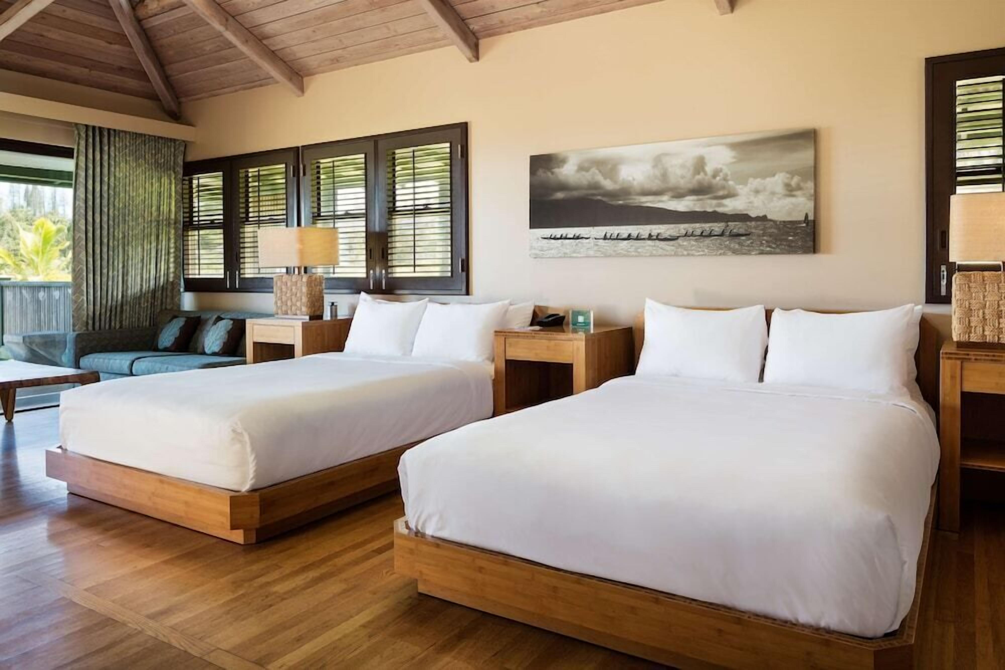 A hotel room with two beds, a sofa, a wooden ceiling, and a large window with blinds.