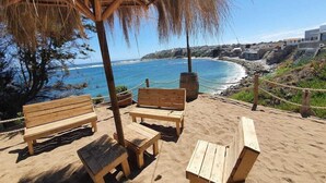 Un espace aménagé avec des bancs et des tables en bois, situé au bord de la plage et offrant une vue sur la côte, avec une eau d’un bleu limpide et un rivage rocheux.