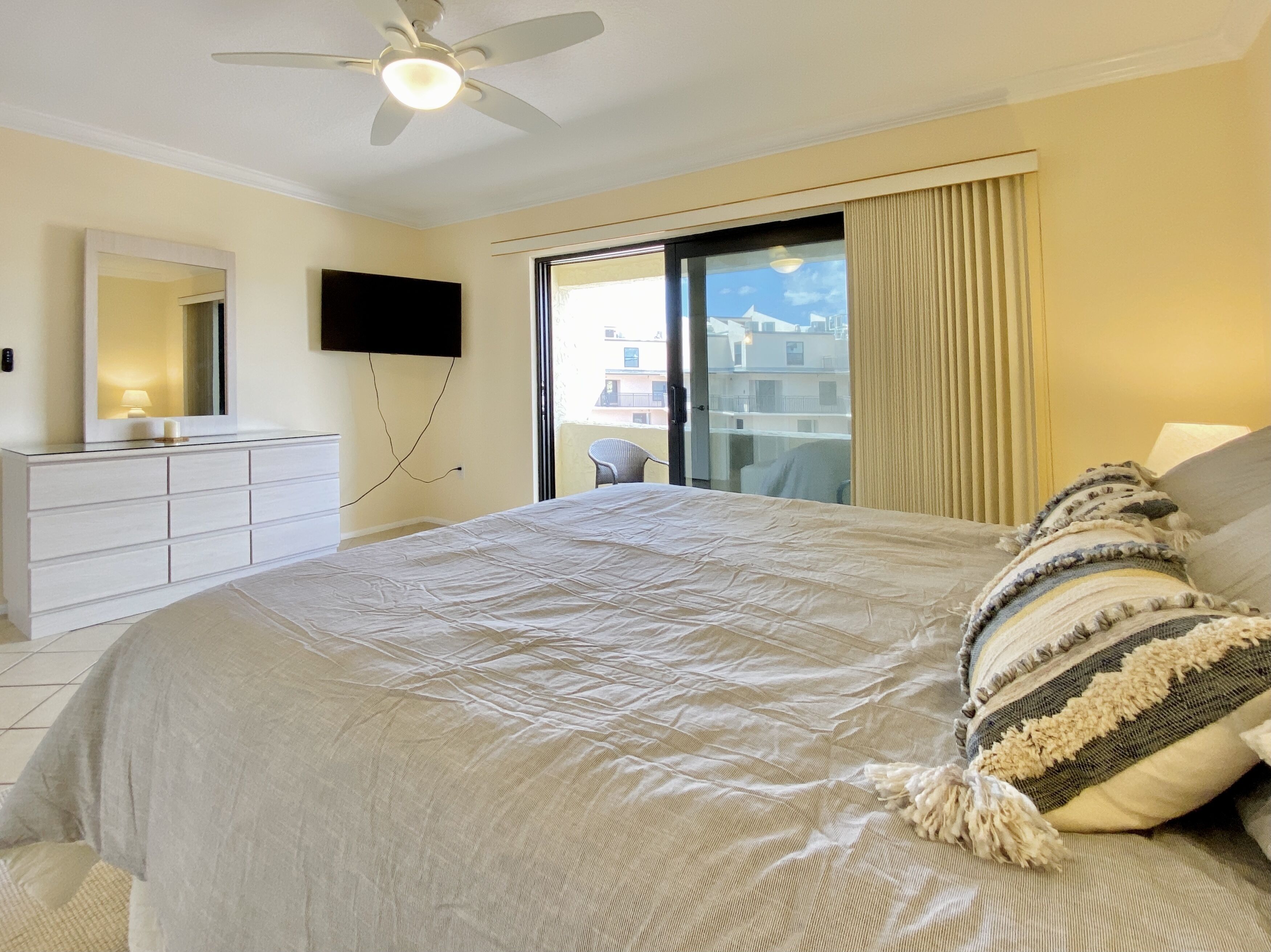 A hotel bedroom with a large bed, a ceiling fan, a TV mounted on the wall, and a balcony with a view of buildings.