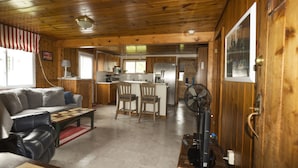Een cabin interieur met een woonkamer, keuken en een eethoek, met houten wanden en vloeren, een bank, stoelen en een ventilator.