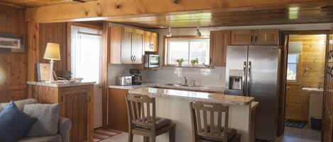 A wooden cabin interior with a kitchen featuring a refrigerator, microwave, and coffee maker. There is a wooden dining table with chairs and a sofa with cushions.