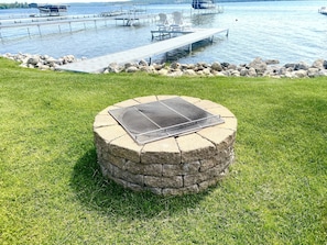 A stone fire pit with a metal grate in a grassy area near a dock and water.