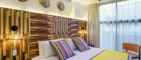 A hotel room with a wooden headboard, a bed with striped pillows, a yellow blanket, a ceiling fan, and a view of the city through large windows.