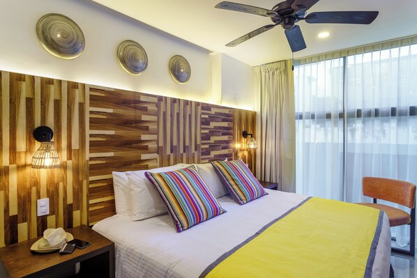 A hotel room with a wooden headboard, a bed with striped pillows, a yellow blanket, a ceiling fan, and a view of the city through large windows.
