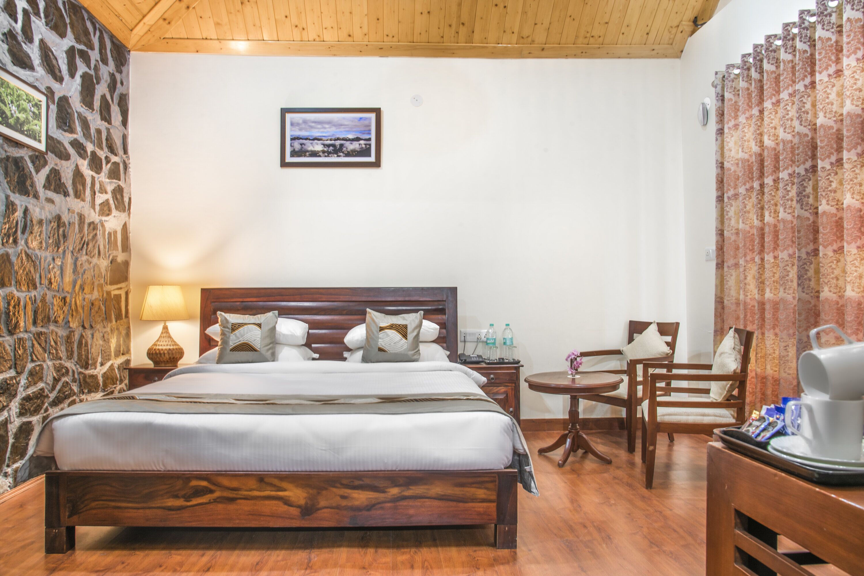 Hotelkamer met een houten bed, een stenen muur, een lijst met een foto aan de muur, en een kleine tafel met stoelen.