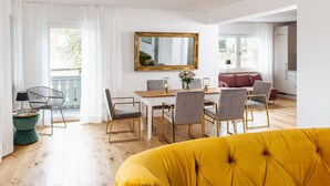 Een lichte, moderne woonkamer met een houten eettafel, gestoffeerde grijze stoelen, een gele bank en een grote spiegel aan de muur.